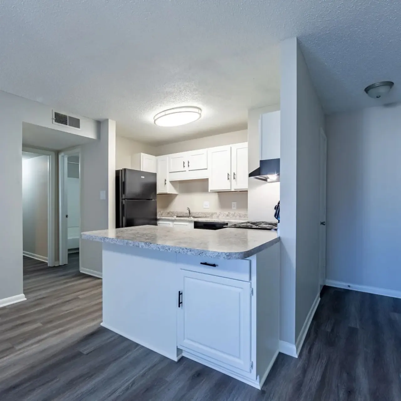 A modern kitchen featuring white cabinets and a black refrigerator, with a countertop providing a view to the adjacent living space. The flooring is a light-colored laminate and the walls are painted in neutral tones.