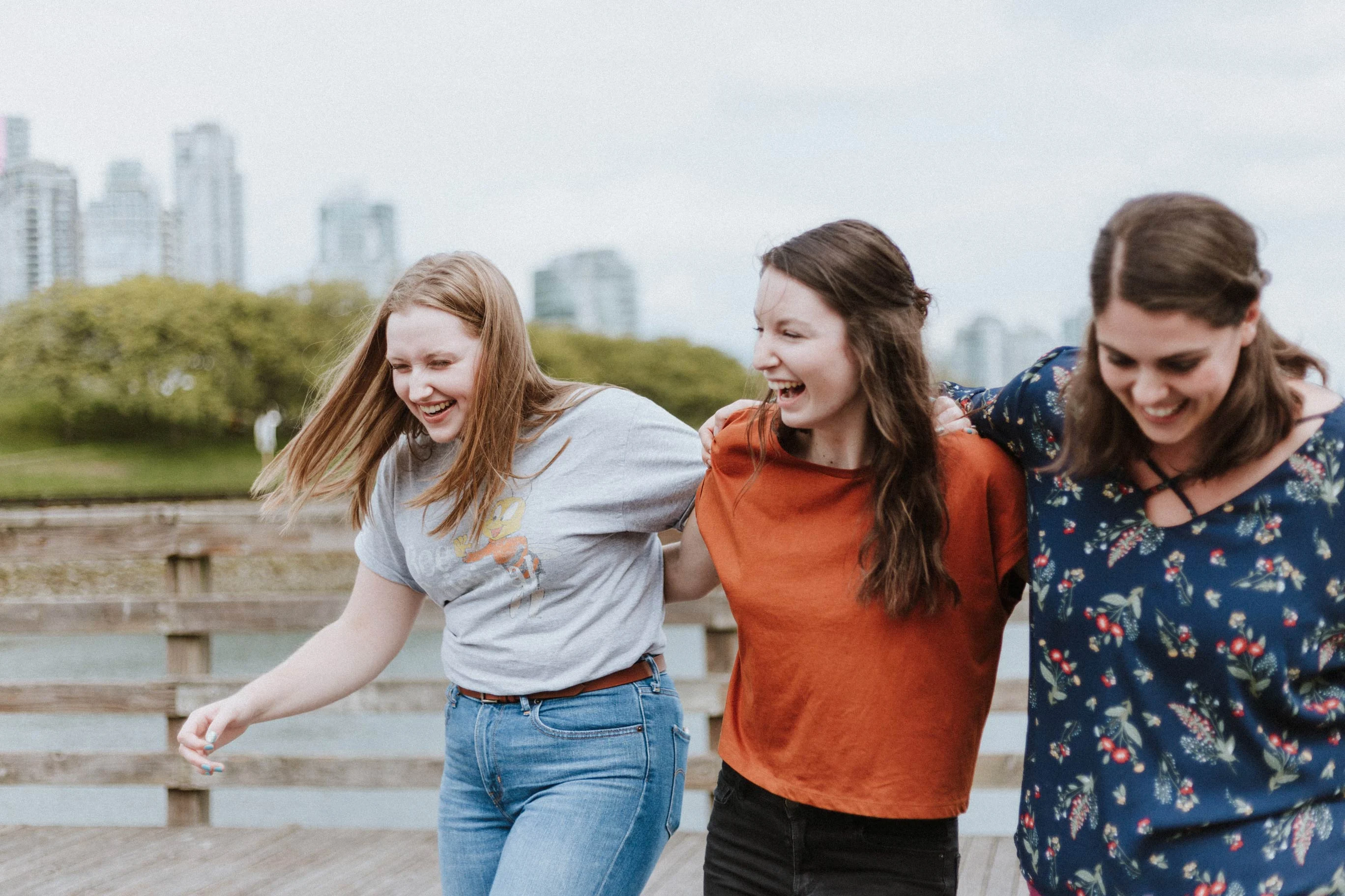 Three young women laughing together while walking along a wooden pier with a cityscape in the background.