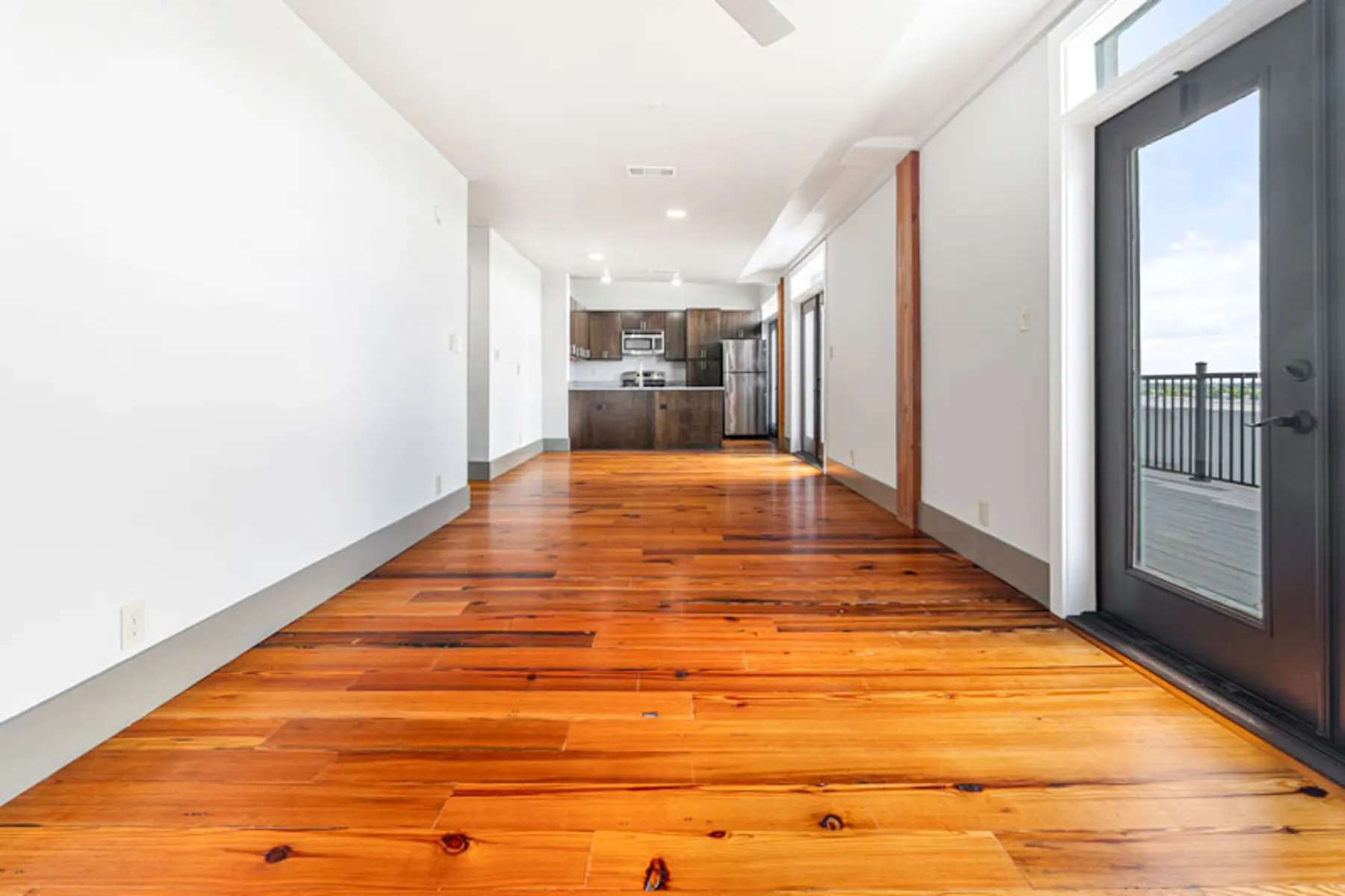 A spacious, empty living area with wooden flooring and modern kitchen visible in the background, featuring large windows and a door leading to a balcony.