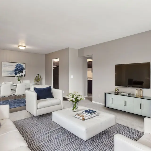 A modern living room featuring white sofas with blue cushions, a coffee table in the center, a television mounted on the wall, and a light-colored rug. A dining area is visible in the background along with stylish decor.