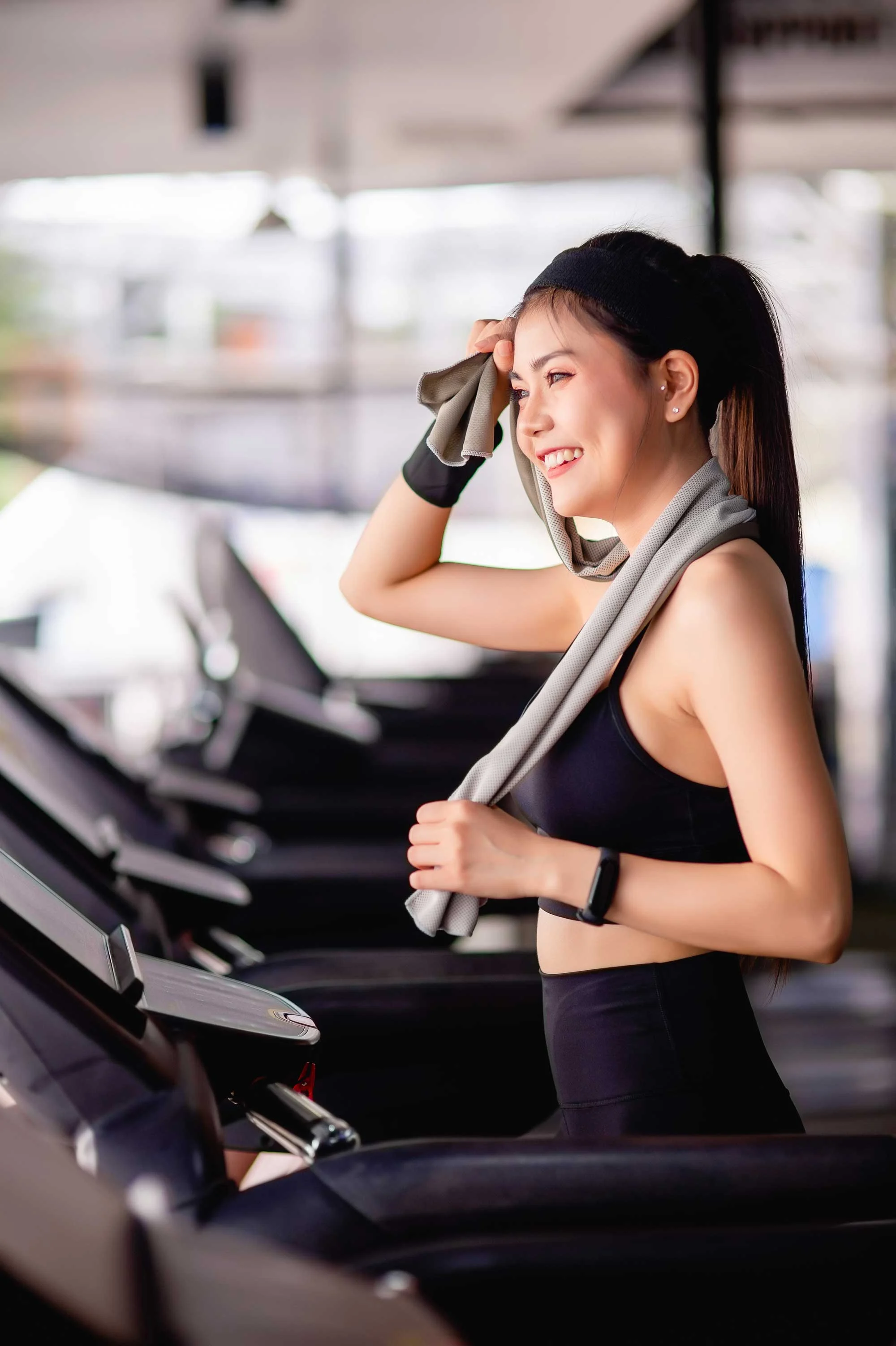 Smiling Woman in Gym A young woman in workout attire smiling while resting on a treadmill in a gym, holding a towel.