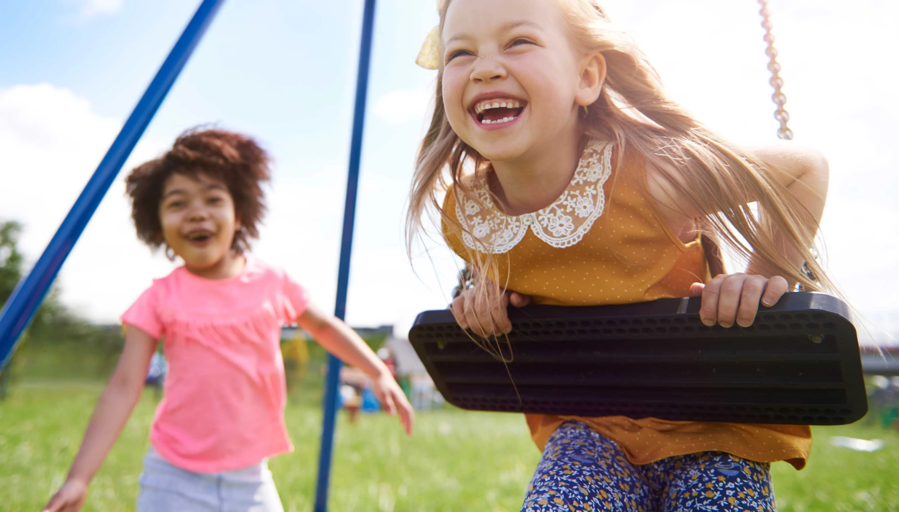 Two young girls are playing on a swing and running in a park. One girl on the swing is laughing joyfully while wearing an orange top with a collar and patterned blue pants, and the other girl, wearing a pink shirt, is running beside her.