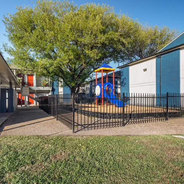 An outdoor playground area enclosed by a black fence, surrounded by buildings and a grassy area.