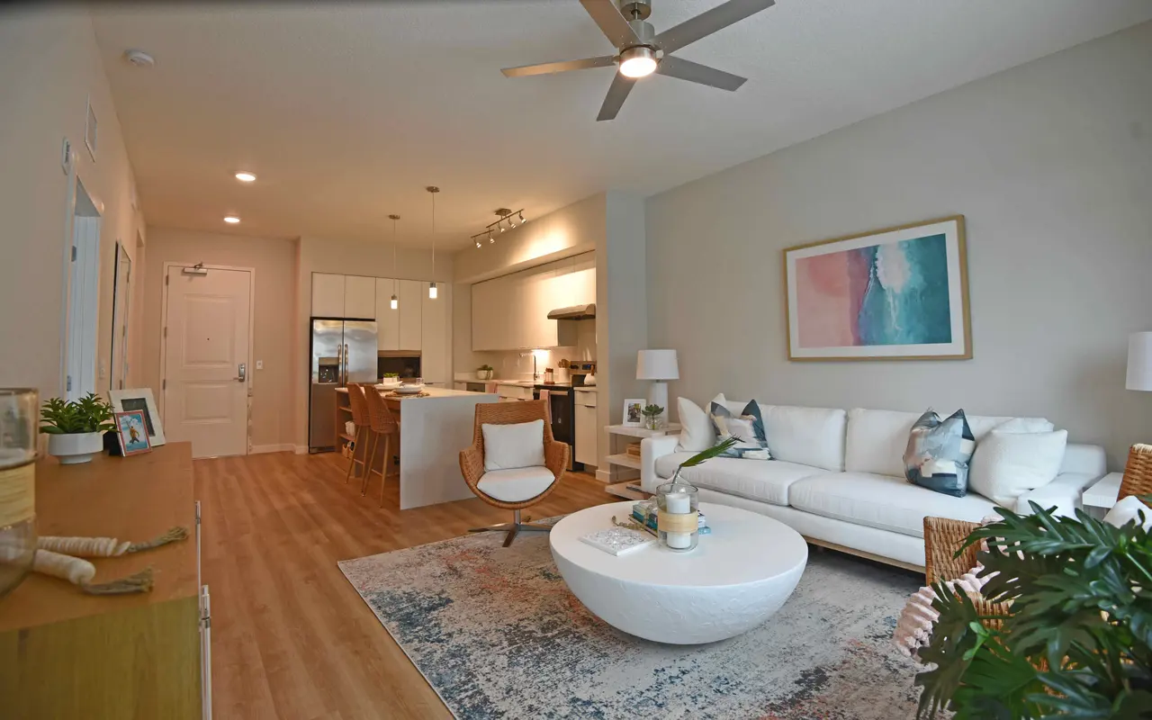 A modern living room with a white couch, a round coffee table, and large windows. There is a dining area in the background with wooden chairs and a kitchen space featuring stainless steel appliances. The room has a light color scheme and decorative plants.