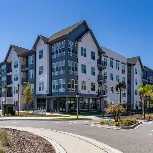 A modern apartment complex with multiple stories, featuring a mix of white and grey exteriors, large windows, and balconies. The setting includes manicured landscaping and palm trees, with a clear blue sky above.