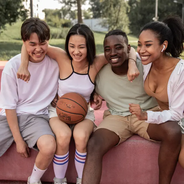A diverse group of four young friends sits together on a pink surface, smiling and holding a basketball, enjoying their time outdoors.