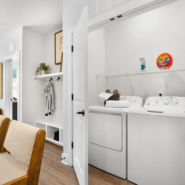 A view of a laundry room within a modern living space. The laundry area features a washer and dryer, and is separated by a white door. The main area has a wooden dining table and beige chairs, with artwork on the walls and large windows allowing natural light.