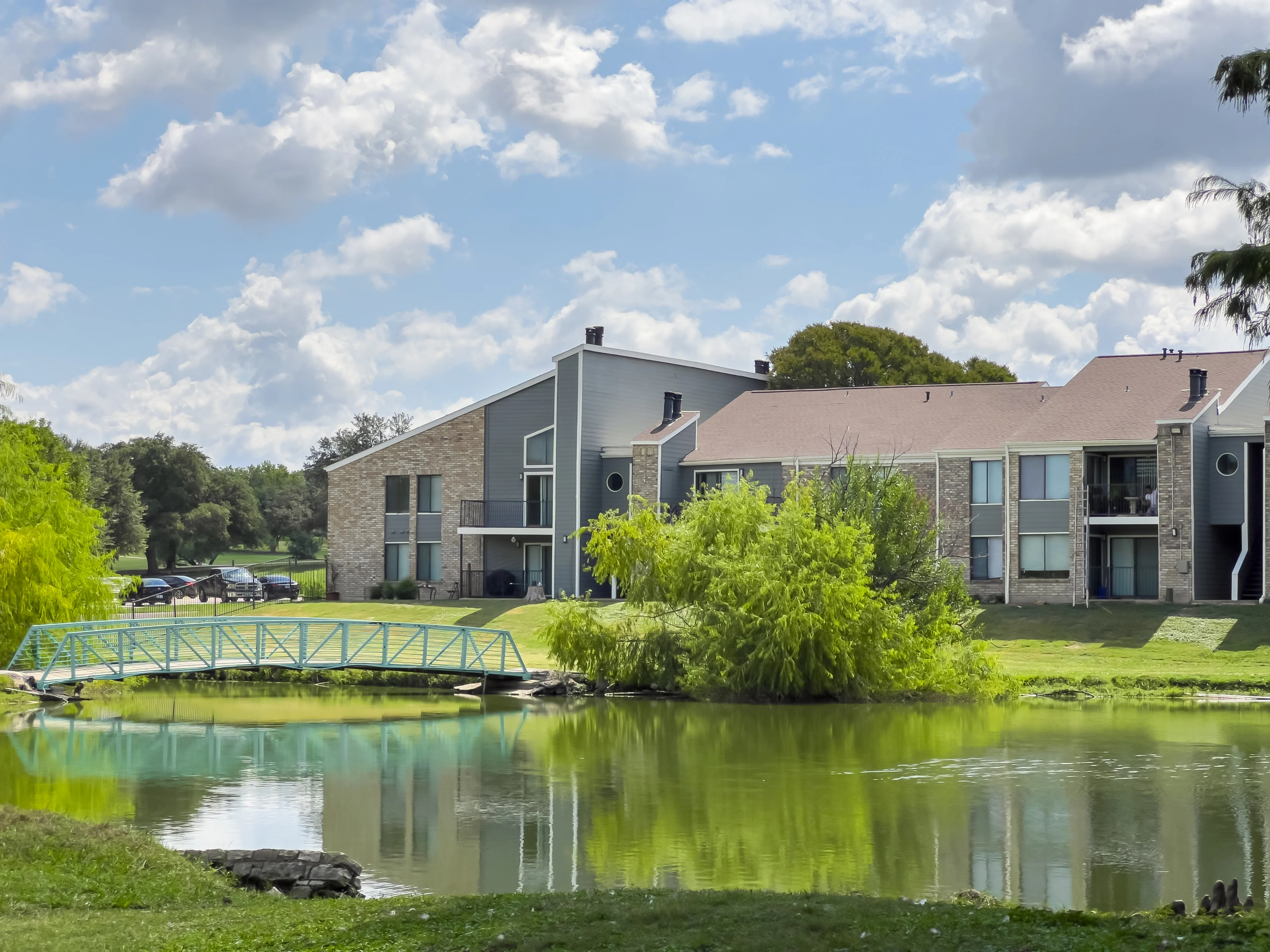 Lakefront Apartment Complex A scenic view of an apartment complex beside a calm lake, featuring green grass, a small blue footbridge, and trees with fresh foliage under a partly cloudy sky.