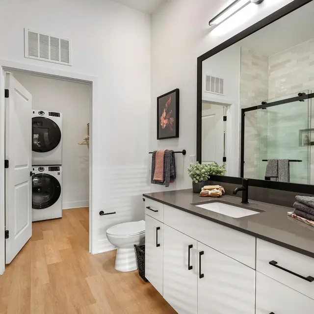 A modern bathroom featuring a spacious vanity with a dark countertop, a large mirror, and stylish storage drawers. The room includes a separate toilet area and a glass shower enclosure. A pair of washing machines are visible in an alcove, adding convenience to the space.
