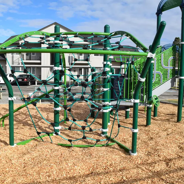 Playground Climbing Structure A green climbing structure at a playground, featuring ropes and nets, surrounded by wood chips and a fence.