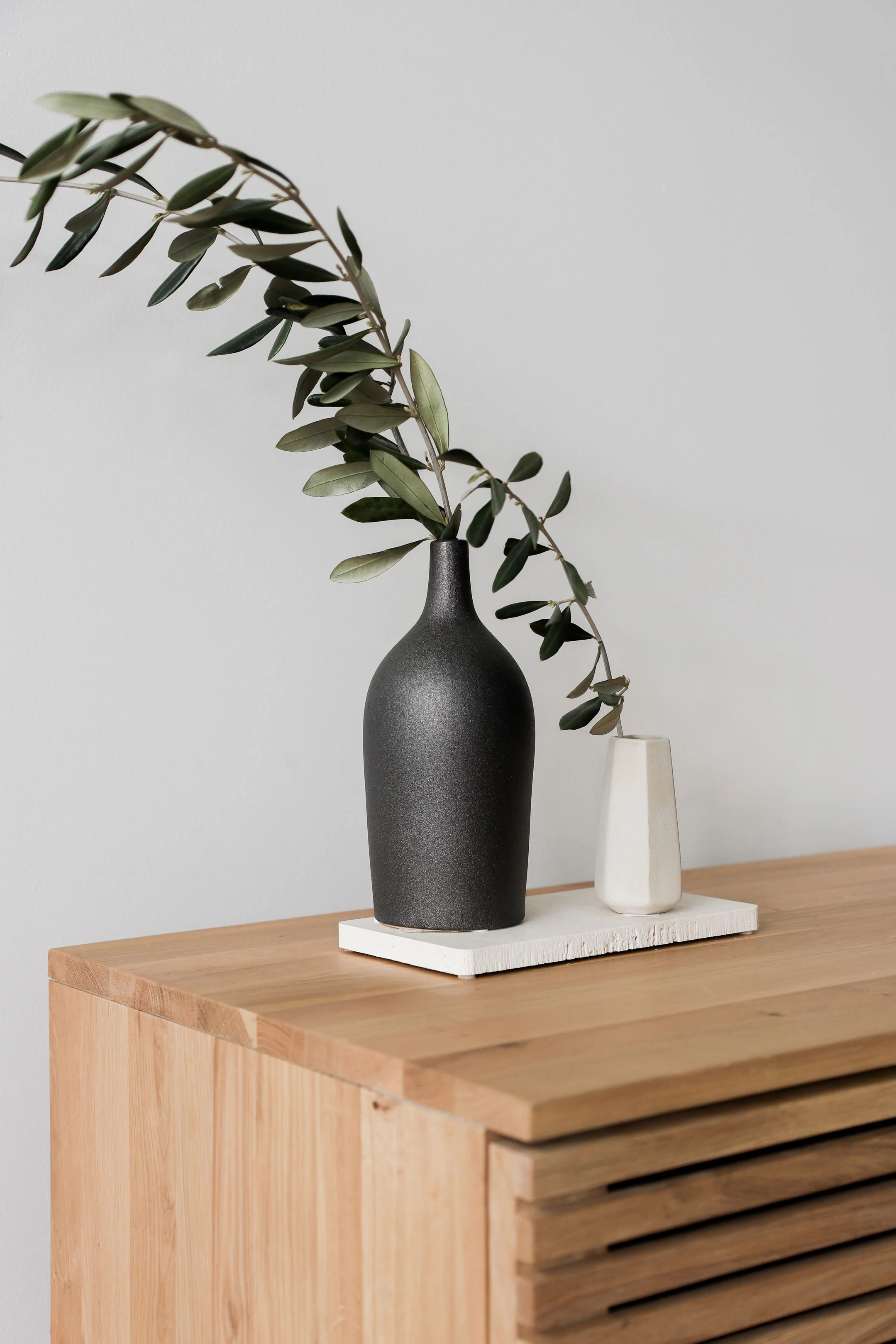 Elegant Vase Arrangement A black ceramic vase with an olive branch is placed on a wooden surface. Next to it is a smaller white vase on a rectangular tray.