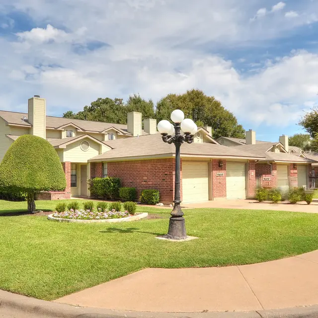 A residential area featuring well-maintained houses and landscaping, with a roundabout in the foreground. A lamp post stands near the center, surrounded by manicured grass and shrubs.