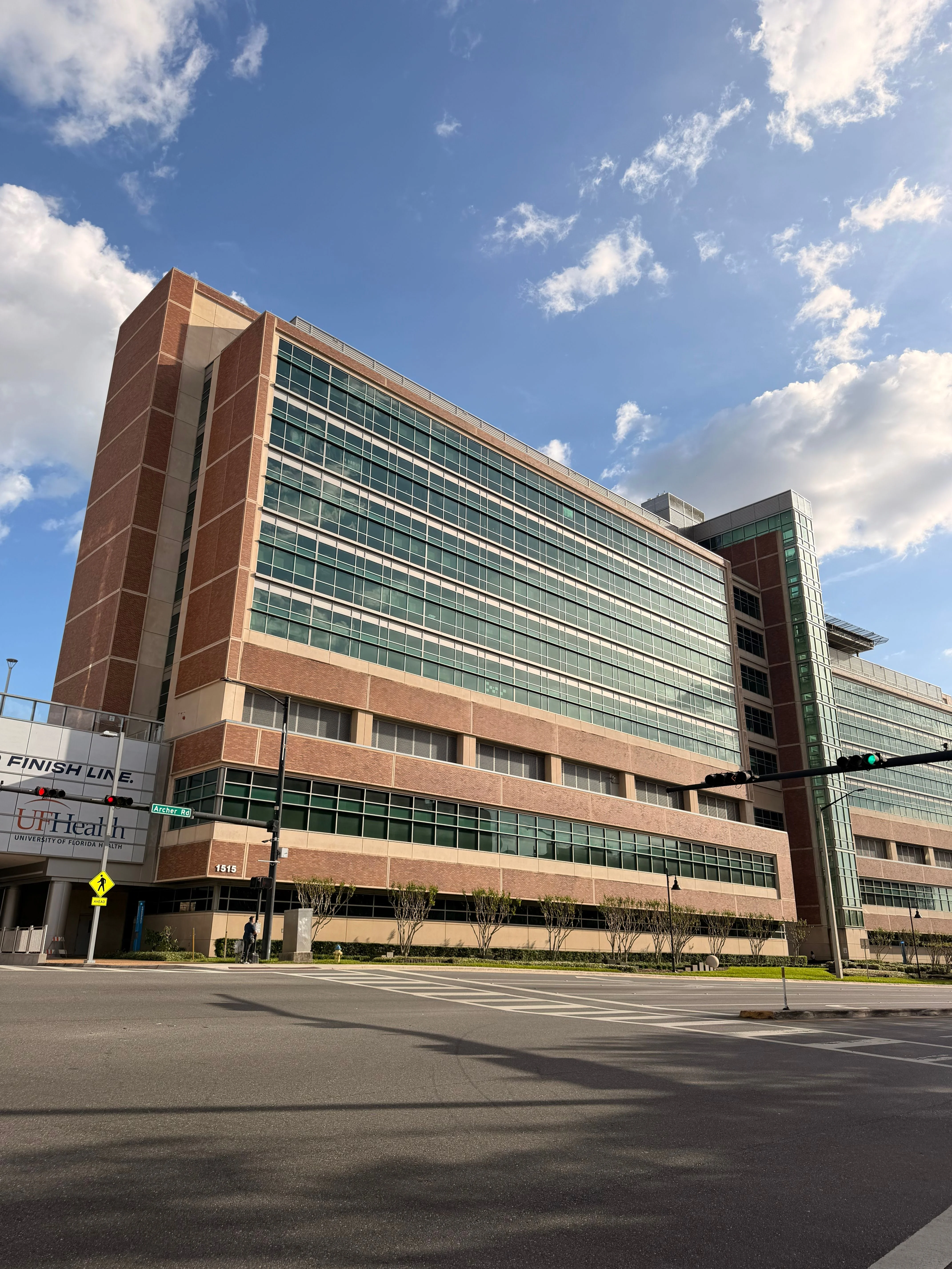 A multi-story healthcare building with a modern architectural style, featuring large glass windows and brick exterior. The sky is partly cloudy with blue sections visible.