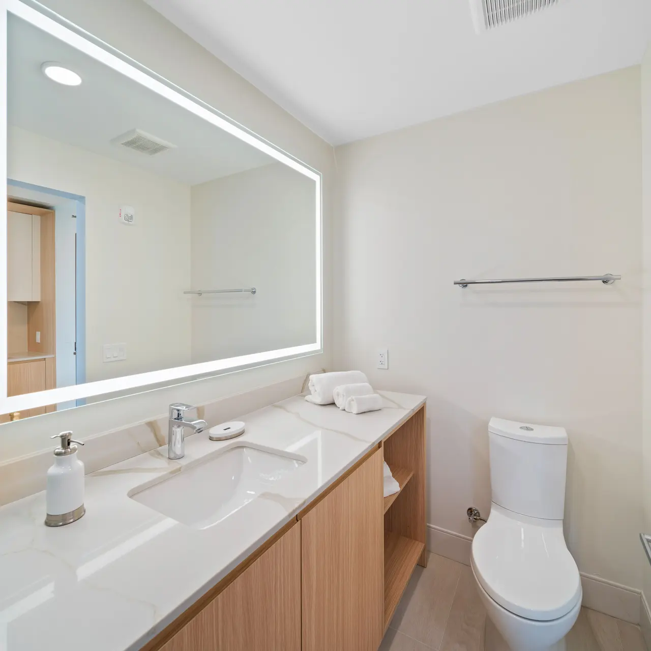 A modern bathroom featuring a stylish illuminated mirror above a sink with a minimalist design. The space has light-colored walls, a wooden vanity, and a white toilet. There is a towel bar on the wall and a warm, inviting atmosphere.