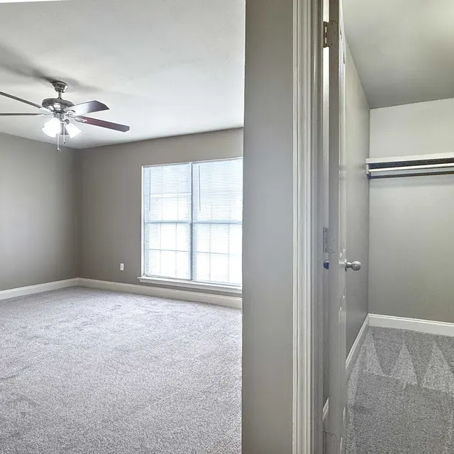 A spacious bedroom featuring a light gray carpet, a ceiling fan, and a large window with white blinds. A closet is visible on the right side of the image.