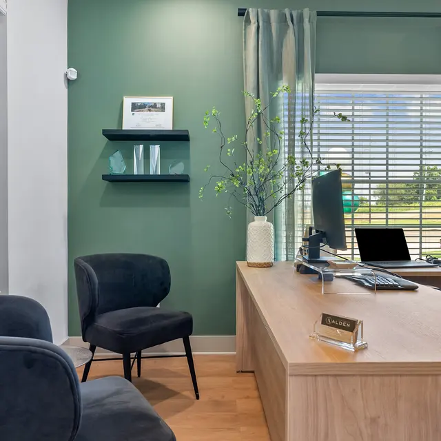 A modern office space featuring a wooden desk, black chairs, and green walls with natural light coming in from a window.