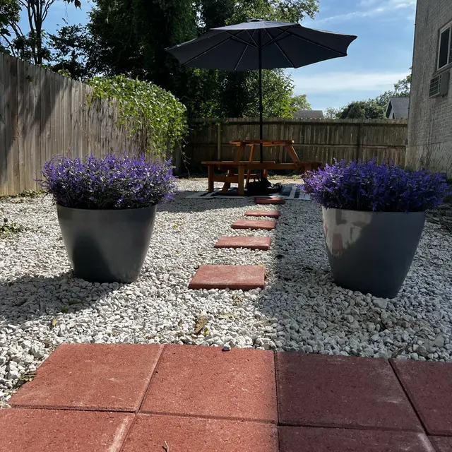 A backyard scene with two large flower pots filled with purple flowers, leading pathway of stepping stones on white gravel, and a wooden bench under a black umbrella.