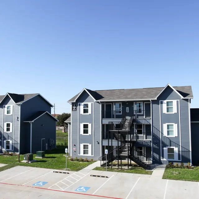 A view of a multi-unit apartment complex featuring several blue-gray buildings with staircases and parking spaces in front on a clear day.