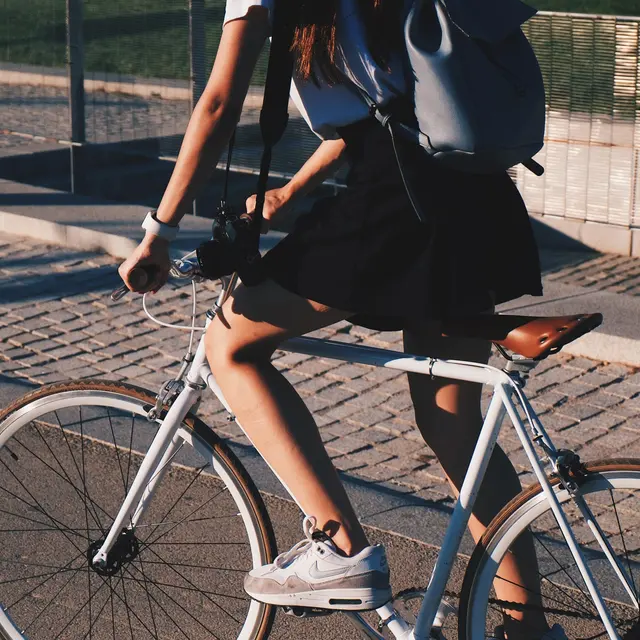 A person in a black skirt and white top rides a bicycle on a paved path, wearing a backpack and sneakers.