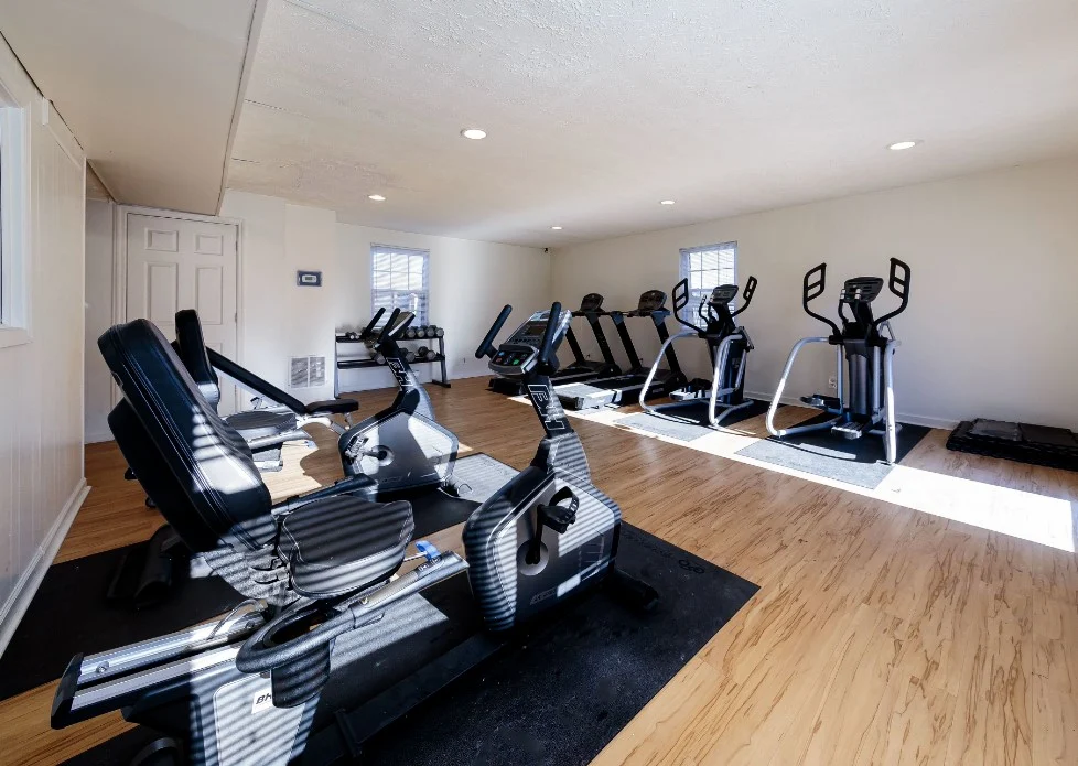 Interior view of a gym filled with various exercise machines including stationary bikes and elliptical trainers on a wooden floor.