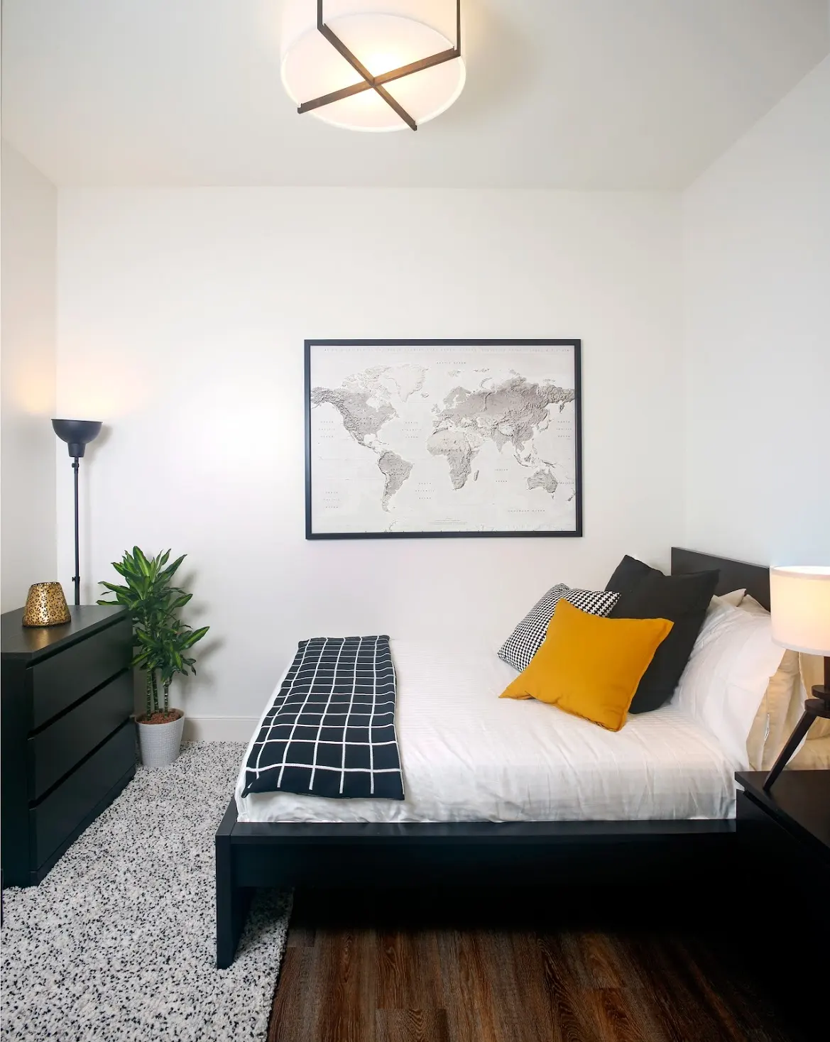 A modern bedroom featuring a bed with white bedding and yellow accent pillow, a world map on the wall, a dresser, and a potted plant.