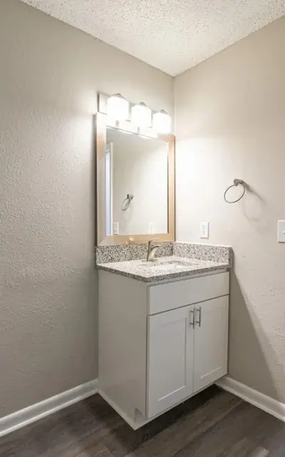 A modern bathroom featuring a small vanity with a granite countertop, a mirror above it, and a well-lit environment. In the background, a toilet can be seen through an open door.