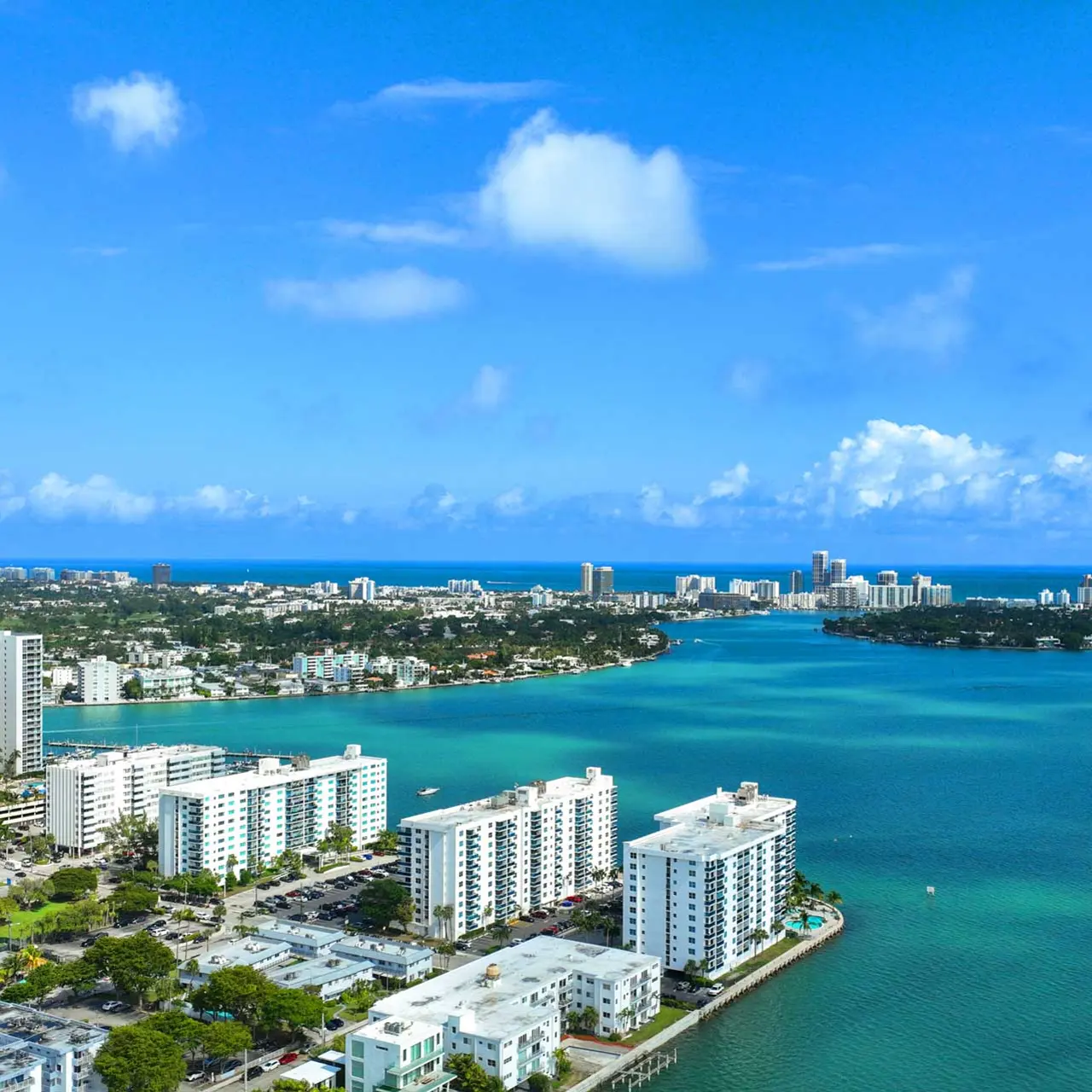 Aerial view of Miami with a vibrant blue ocean and skyline in the background, showcasing buildings along the shore and lush greenery.