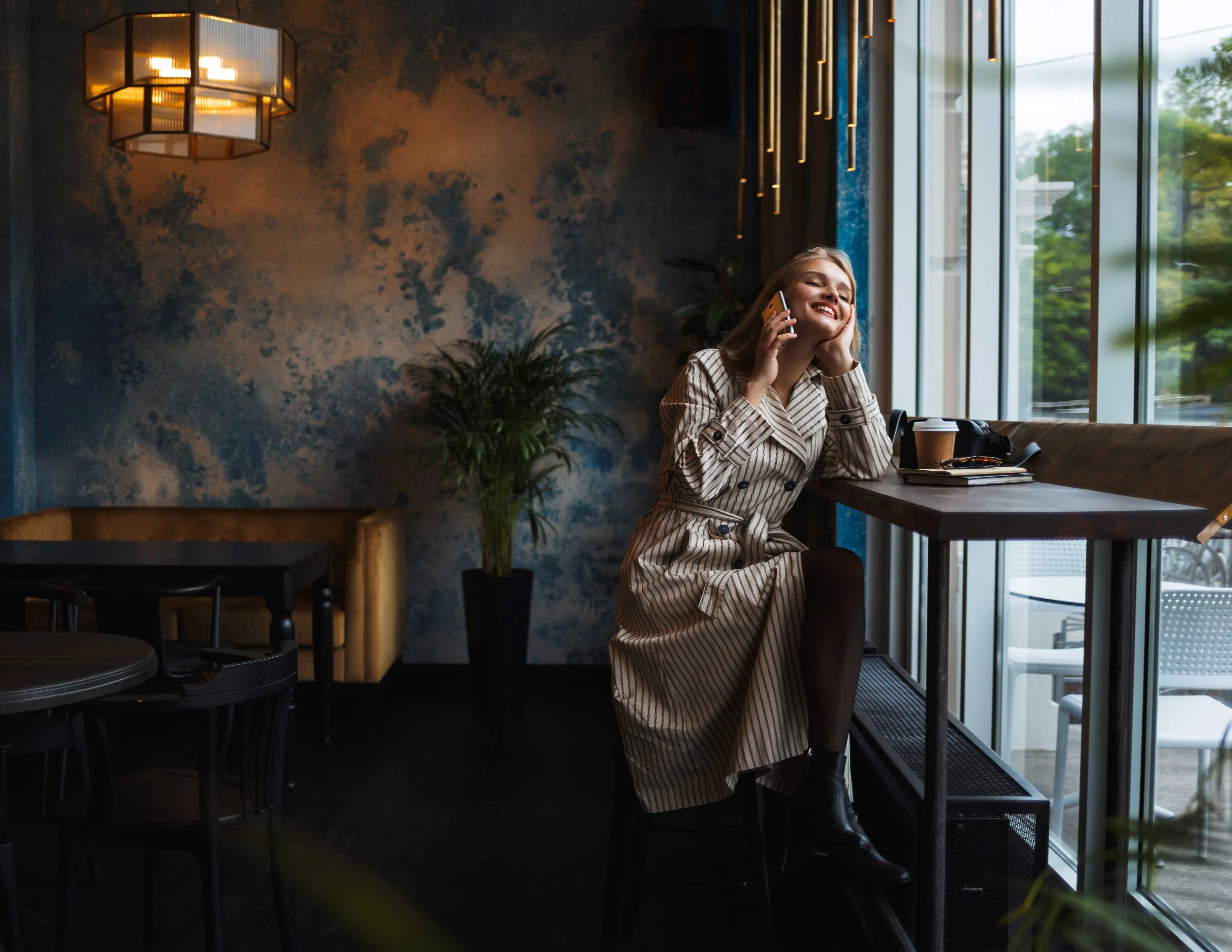 Joyful Moment in a Cafe A woman sitting in a cafe with a striped dress, smiling and holding a phone while sitting near a large window with greenery outside.