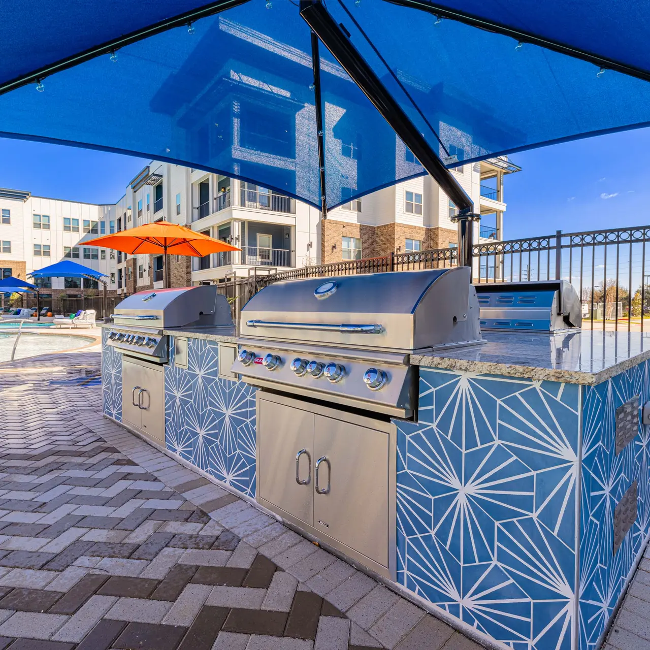 A modern grill area with two stainless steel barbecues under blue canopies, next to a swimming pool and surrounded by a decorative tile wall.