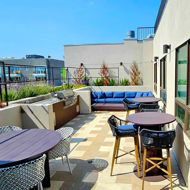 Stylish Rooftop Lounge Area A modern rooftop lounge area with furniture including a blue couch, round tables, and chairs, surrounded by greenery and buildings in the background.