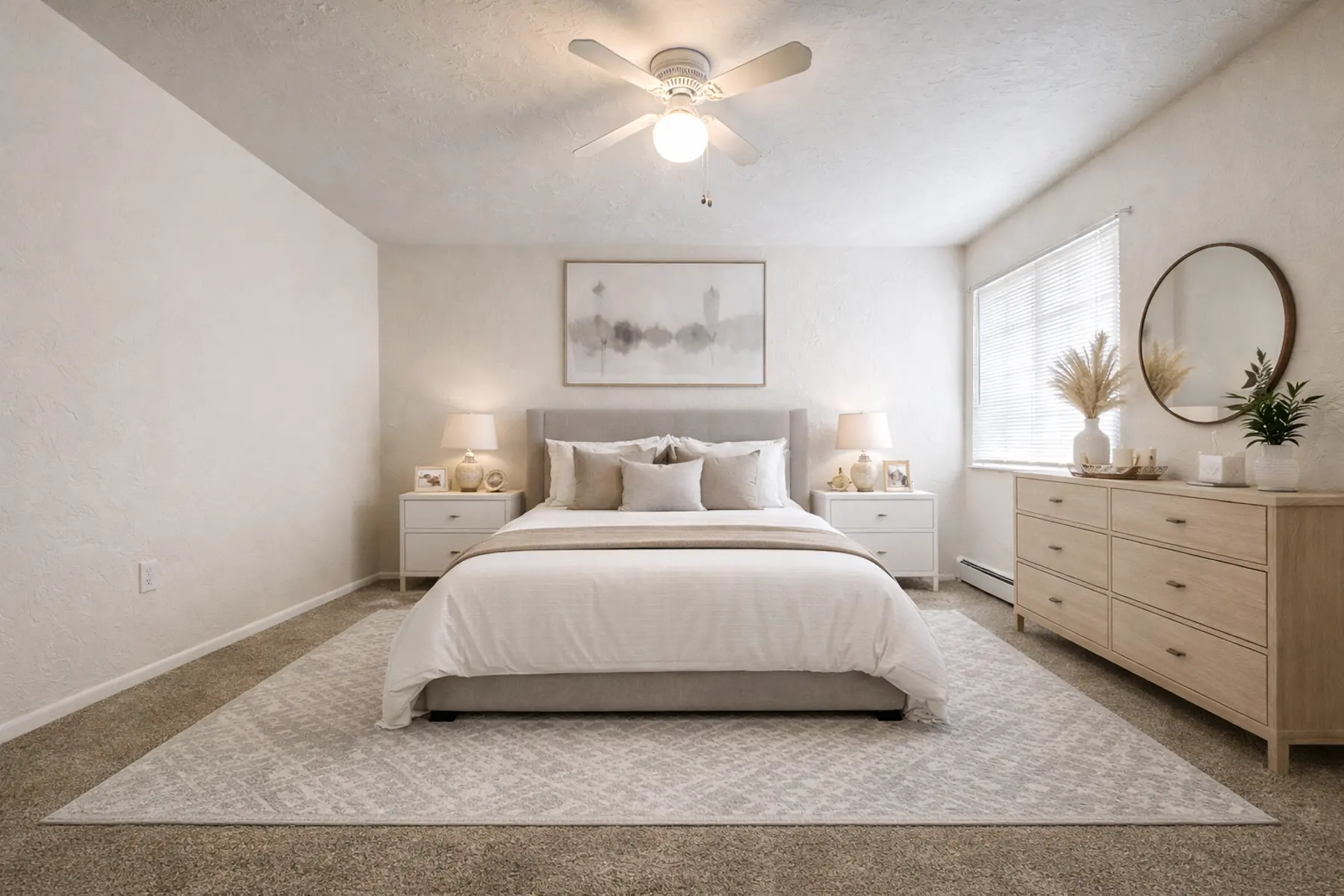 A minimalist bedroom featuring a queen-sized bed with a light gray upholstered headboard, white bedding, and decorative pillows. Two nightstands with lamps are on either side of the bed, and a wooden dresser is against the wall. A large window allows natural light to fill the room, and a round mirror hangs above the dresser.