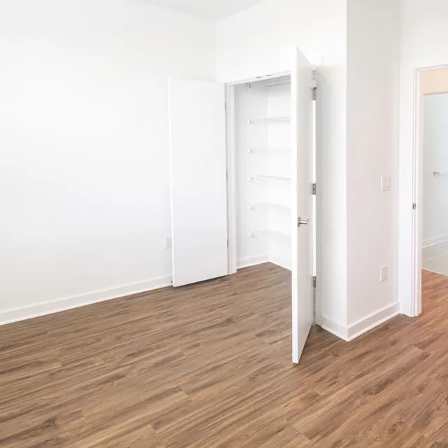 An empty bedroom featuring white walls and wooden flooring, with open doors leading to a closet and a bathroom.