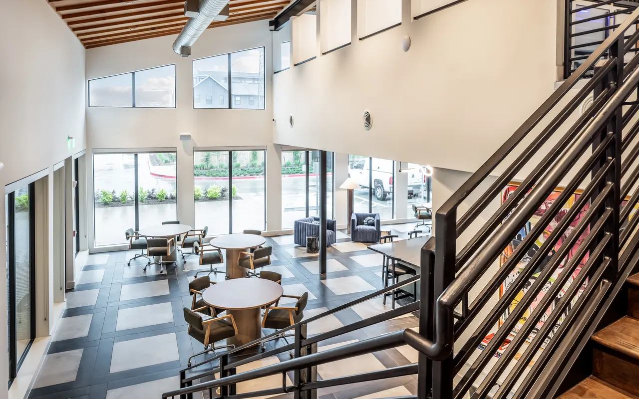 A coworking business center with stairs looking over tables surrounded by chairs, and large windows.