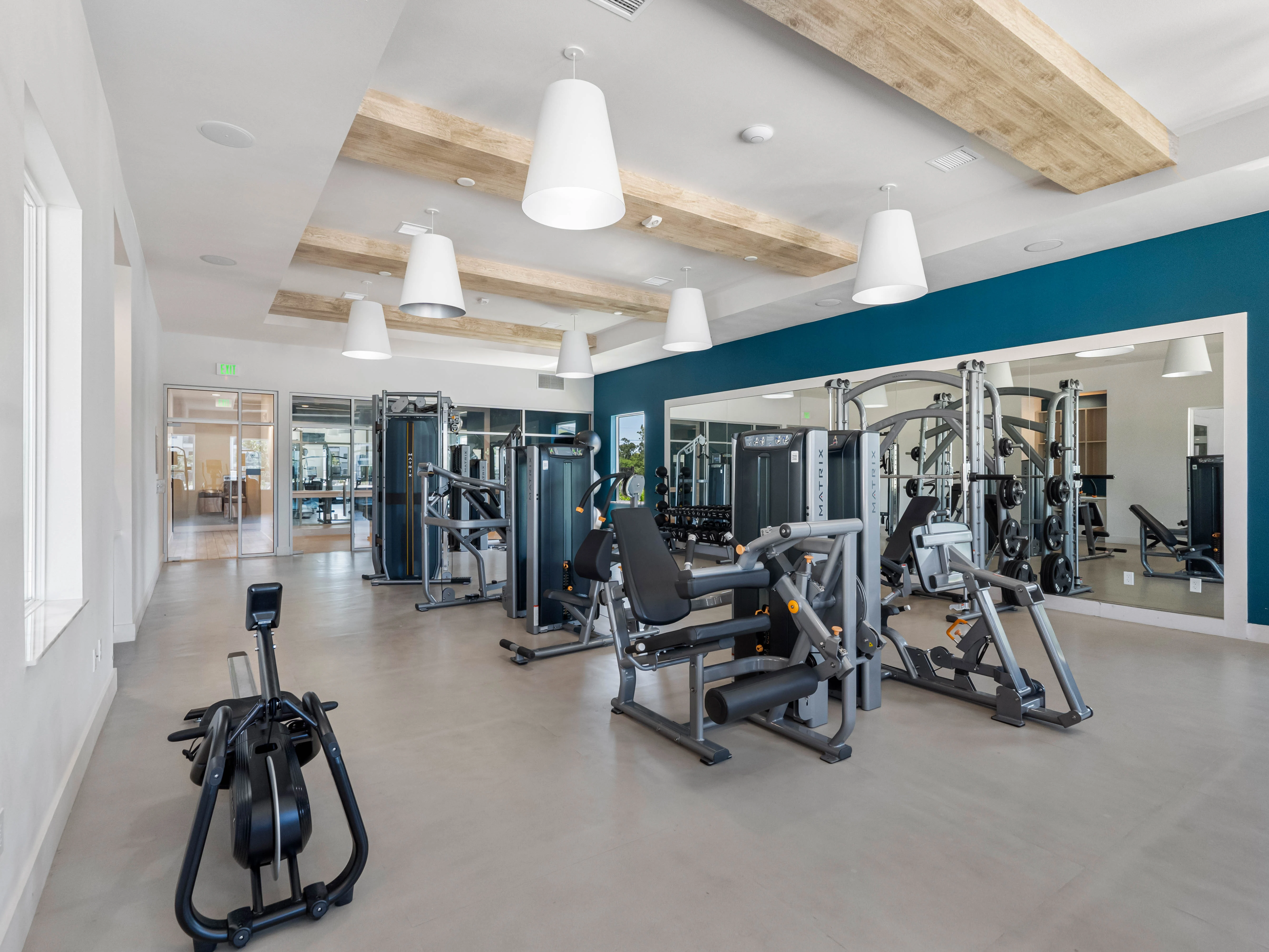 A modern gym interior featuring various exercise machines, bright lighting, and a spacious feel. The design includes light-colored walls with wooden beams on the ceiling and an accent wall in teal.