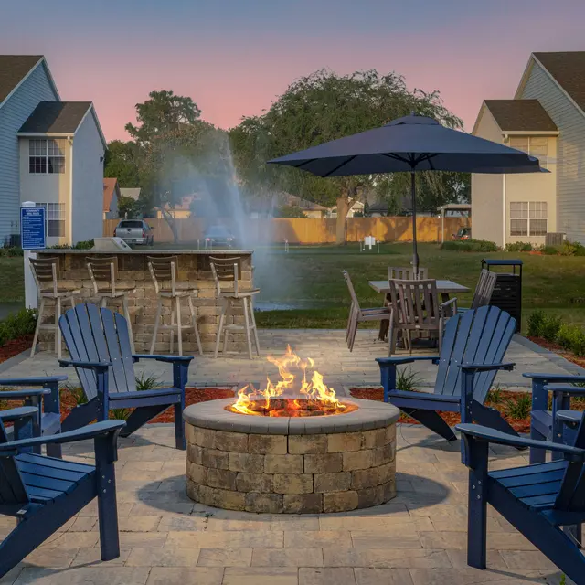 Outdoor patio with fire pit, six Adirondack chairs, an umbrella-shaded table, and a bar with stools, set against a backdrop of houses at sunset.