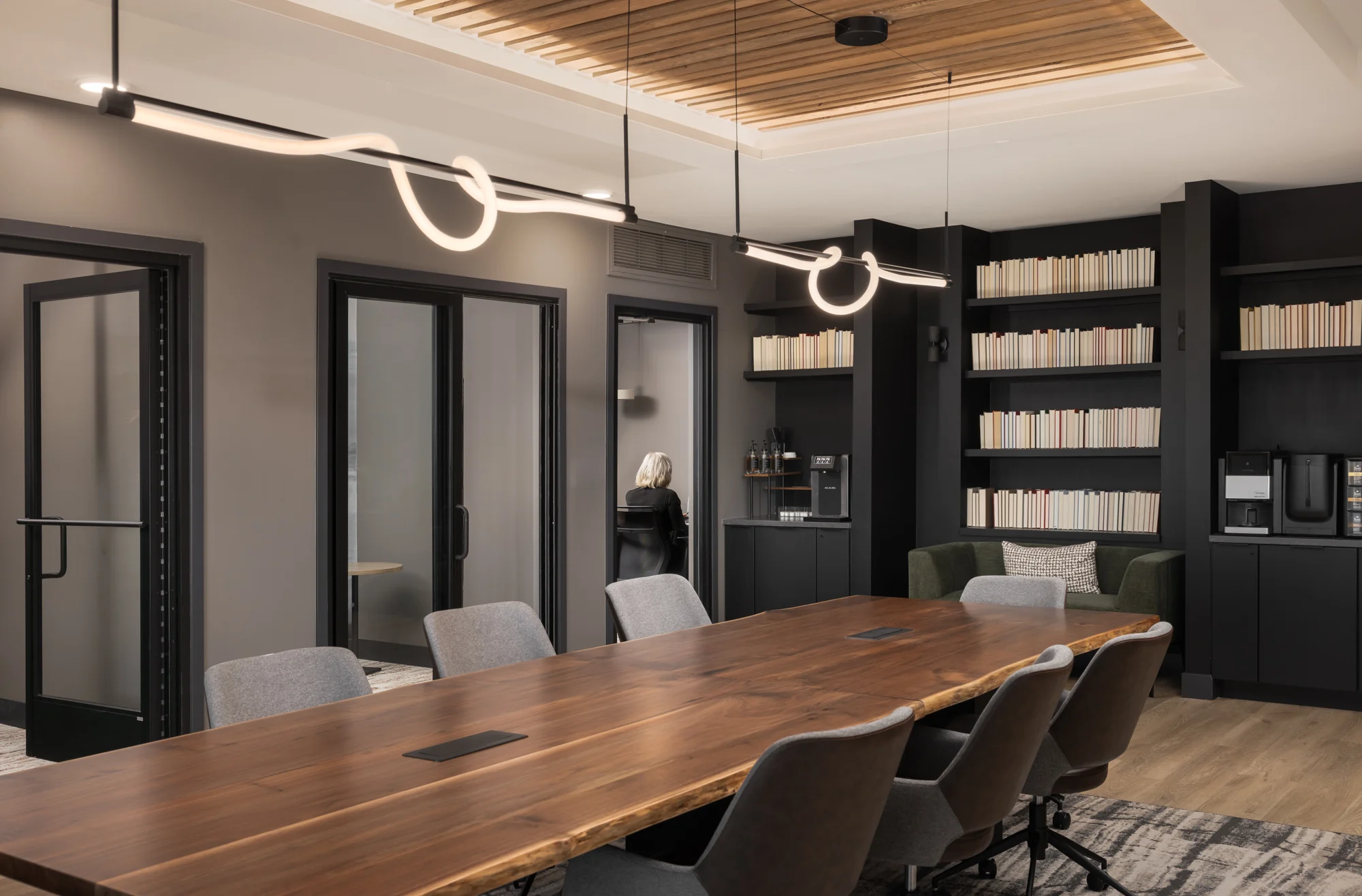 A modern meeting room featuring a long wooden table surrounded by gray chairs, with a sleek light fixture above. On one side, there are large glass doors leading to private rooms, and a bookshelf filled with books on the opposite side.