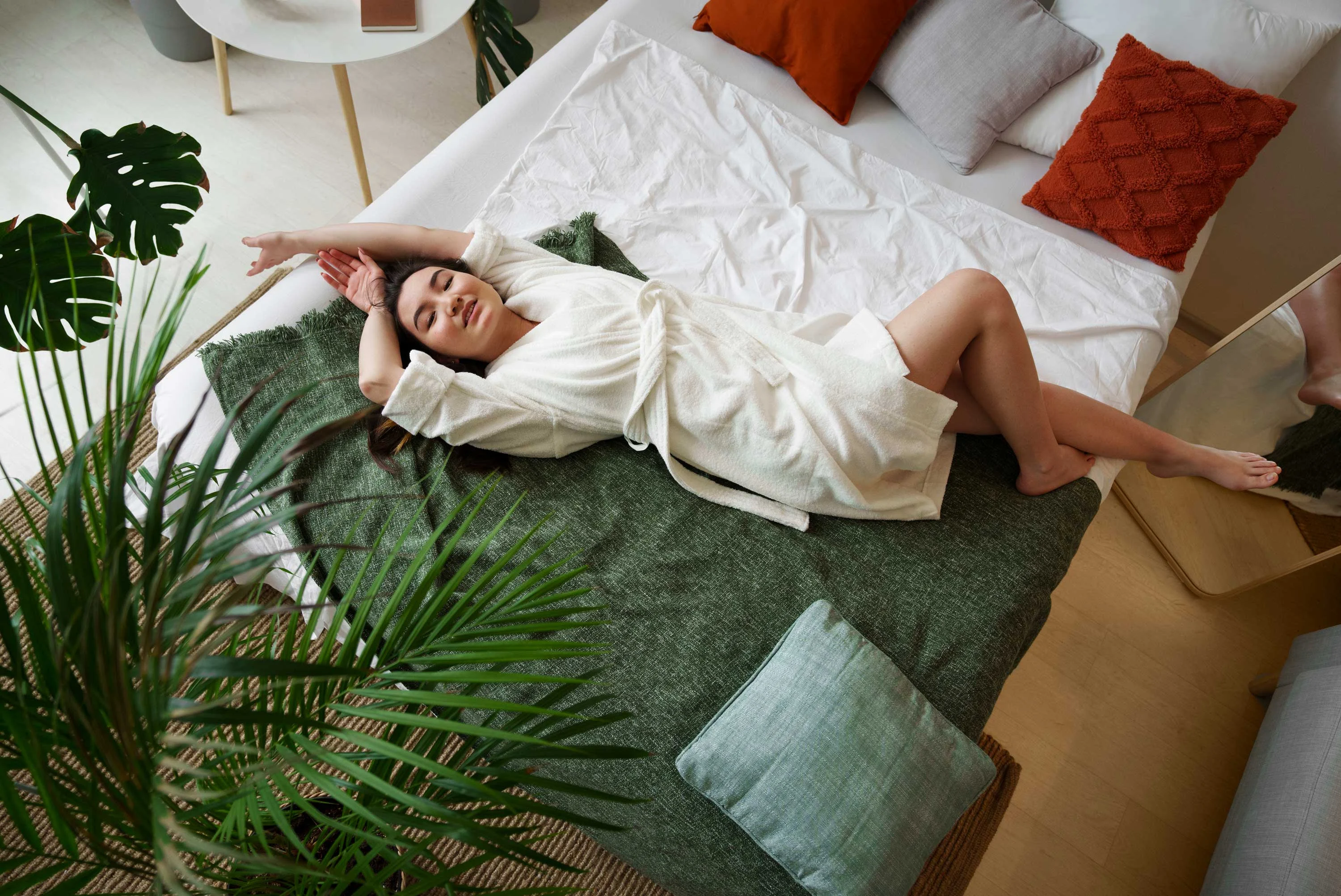The Hideaway A woman in a white robe lying on a bed with green and blue pillows, surrounded by plants and decorative cushions in a modern room.