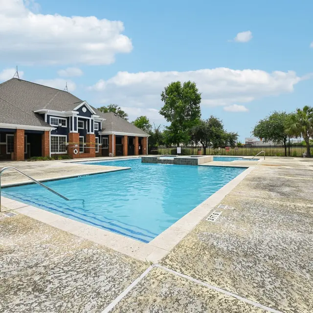 A clean swimming pool surrounded by a textured patio and bordered by tree-filled landscaping.