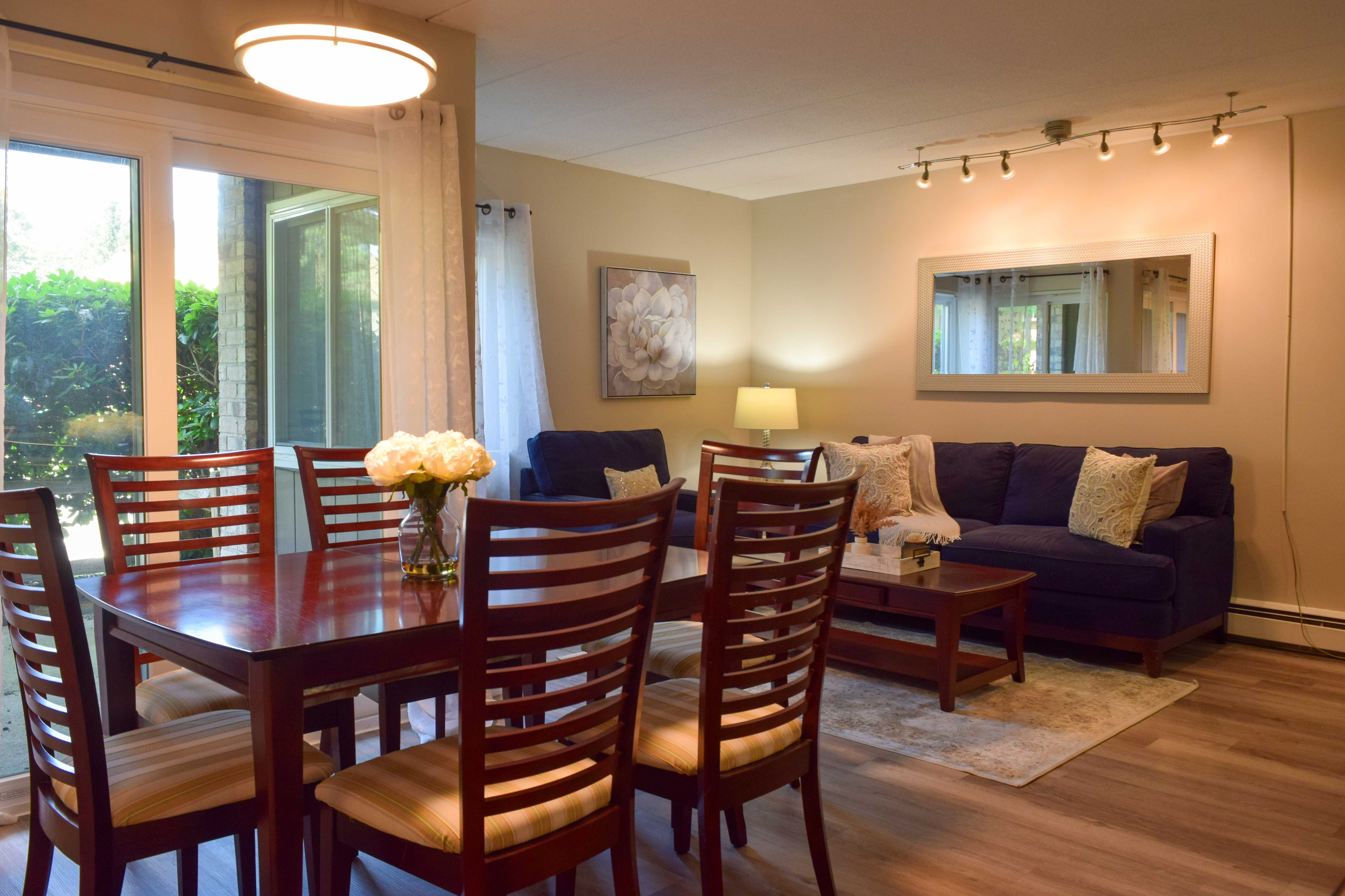 A cozy living and dining area featuring a wooden dining table with chairs and a comfortable blue sofa. Natural light streams in from large windows, and decorative elements like a vase of flowers and wall art are present.