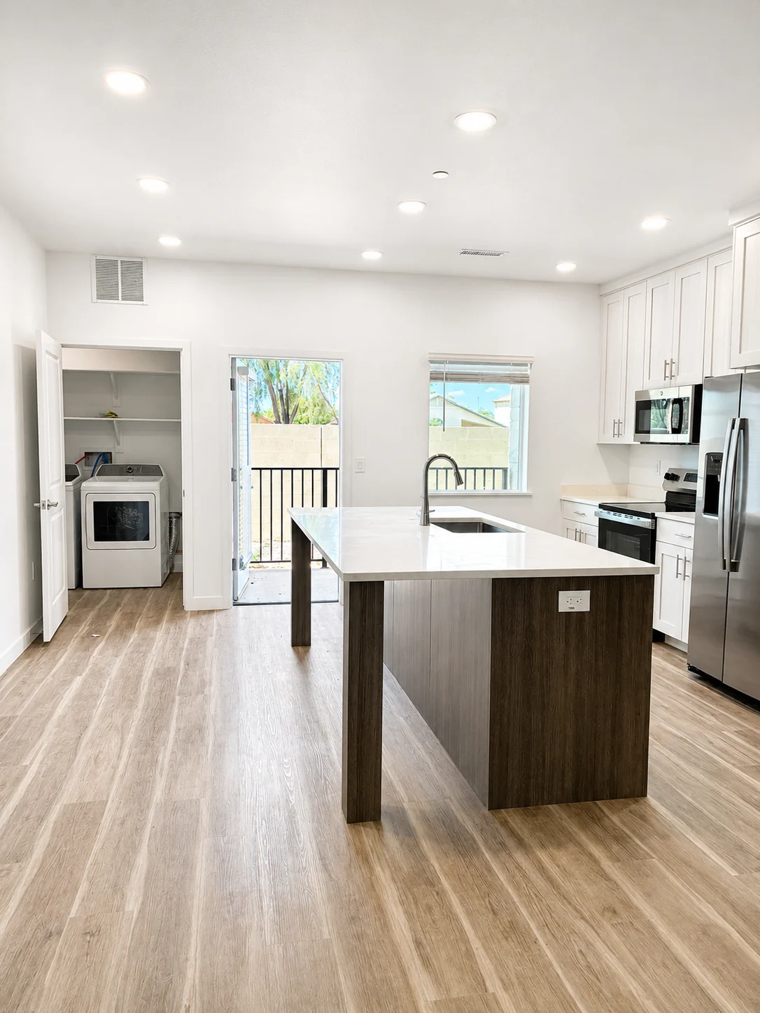 A modern kitchen featuring a large island, stainless steel appliances, and a laundry area. The space has wooden flooring and a door leading to an outdoor balcony.