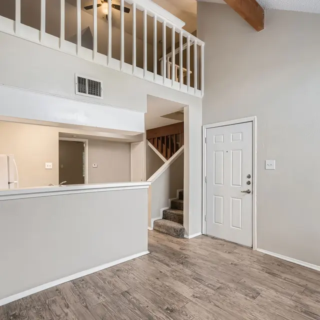 Interior view of a modern living space with a staircase, kitchen area visible, and a front door. Light-colored walls and wooden flooring give a spacious feel.