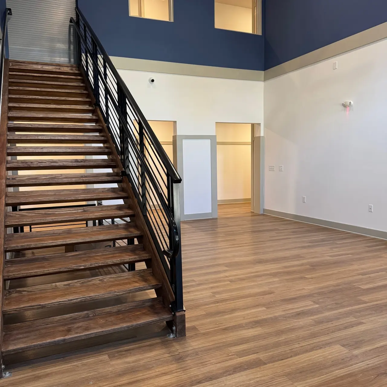A modern indoor space featuring a wooden staircase leading to an upper level, with large windows and a navy blue wall. The room has light wooden flooring and an open layout.