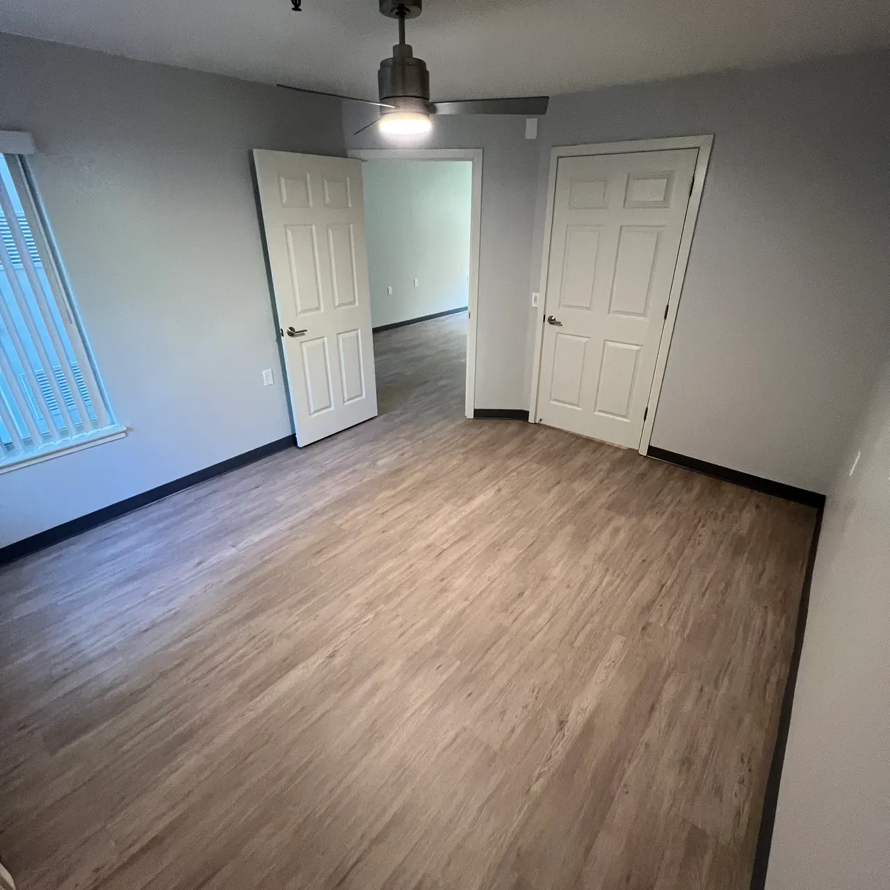 An empty room with a light wood floor, two white doors, and a ceiling light fixture. There are windows with blinds on one side.