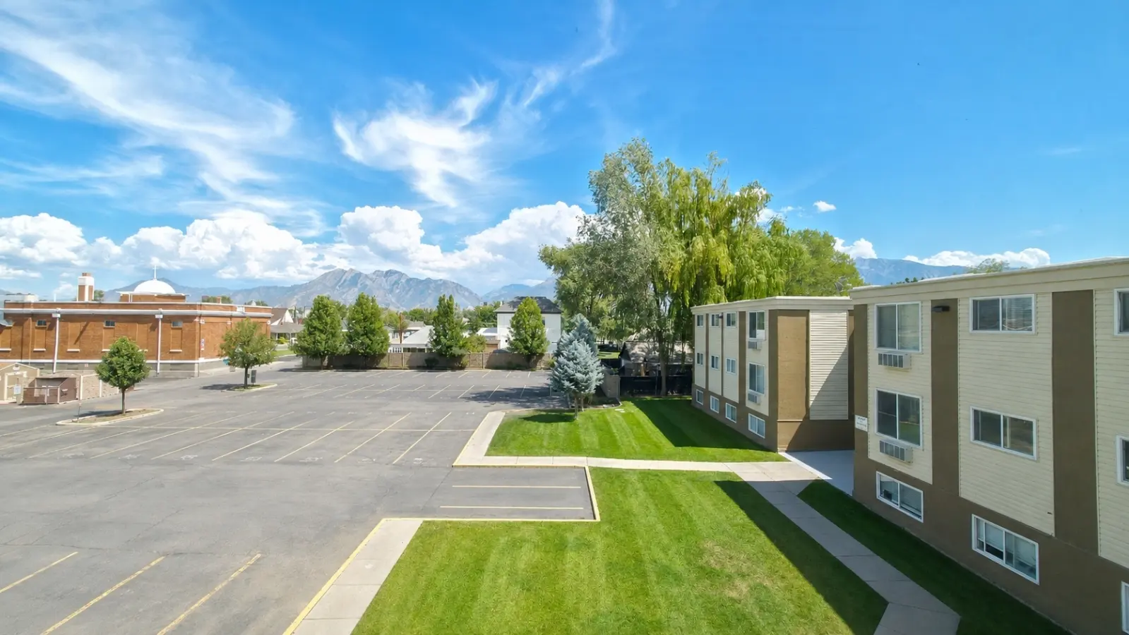 View of an apartment building with a parking lot and green landscaping under a blue sky with clouds.