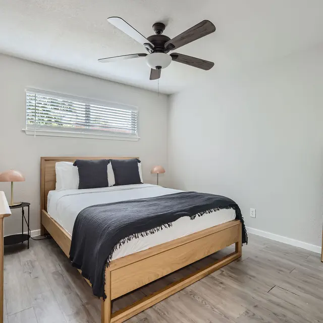 A modern bedroom featuring a wooden bed with a black blanket, two pillows, and a ceiling fan. There is a wooden dresser beside the bed and a large mirror reflecting the room. Natural light filters in through a window with blinds.