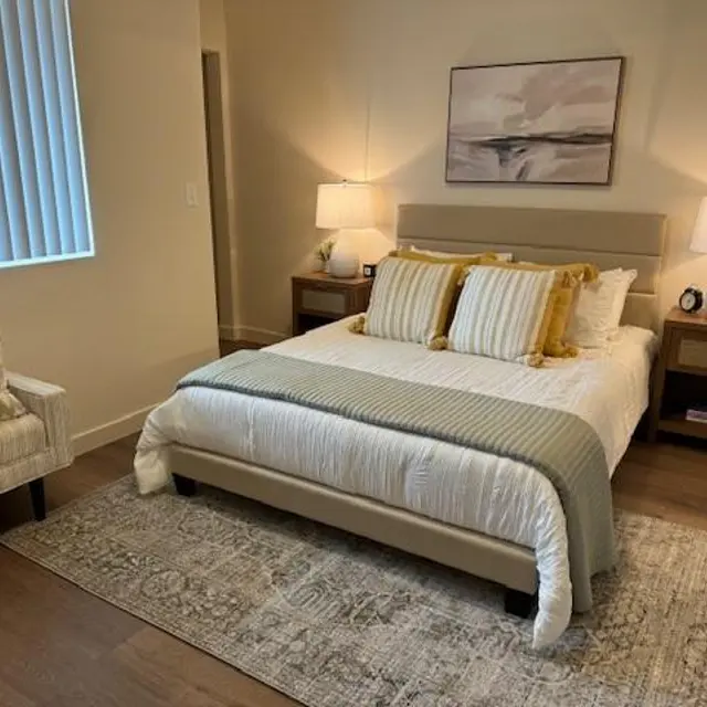 A cozy, well-lit bedroom featuring a comfortable bed with decorative pillows, a light grey blanket, a stylish armchair, and two bedside lamps. The room has wooden flooring and neutral walls, complementing the modern decor.
