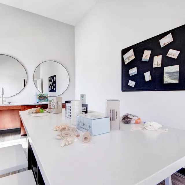 Modern Interior A modern kitchen interior featuring a large white table, a wooden counter with a sink, and two round mirrors on the wall. A bulletin board displays various notes and photos. The floor is made of wood plank. Some kitchen items are scattered across the table.