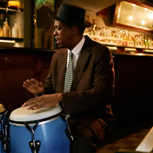 A musician playing conga drums in a bar setting, wearing a brown suit and a hat, with a piano visible on the side.