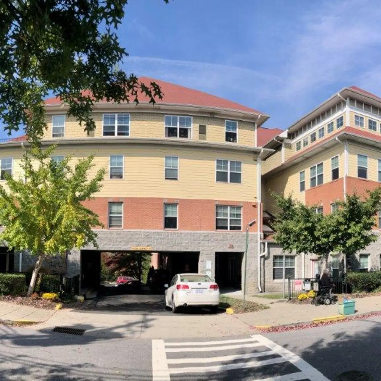 An exterior view of a multi-story building with a combination of brick and siding, showcasing a parking area underneath, surrounded by trees and a clear blue sky.