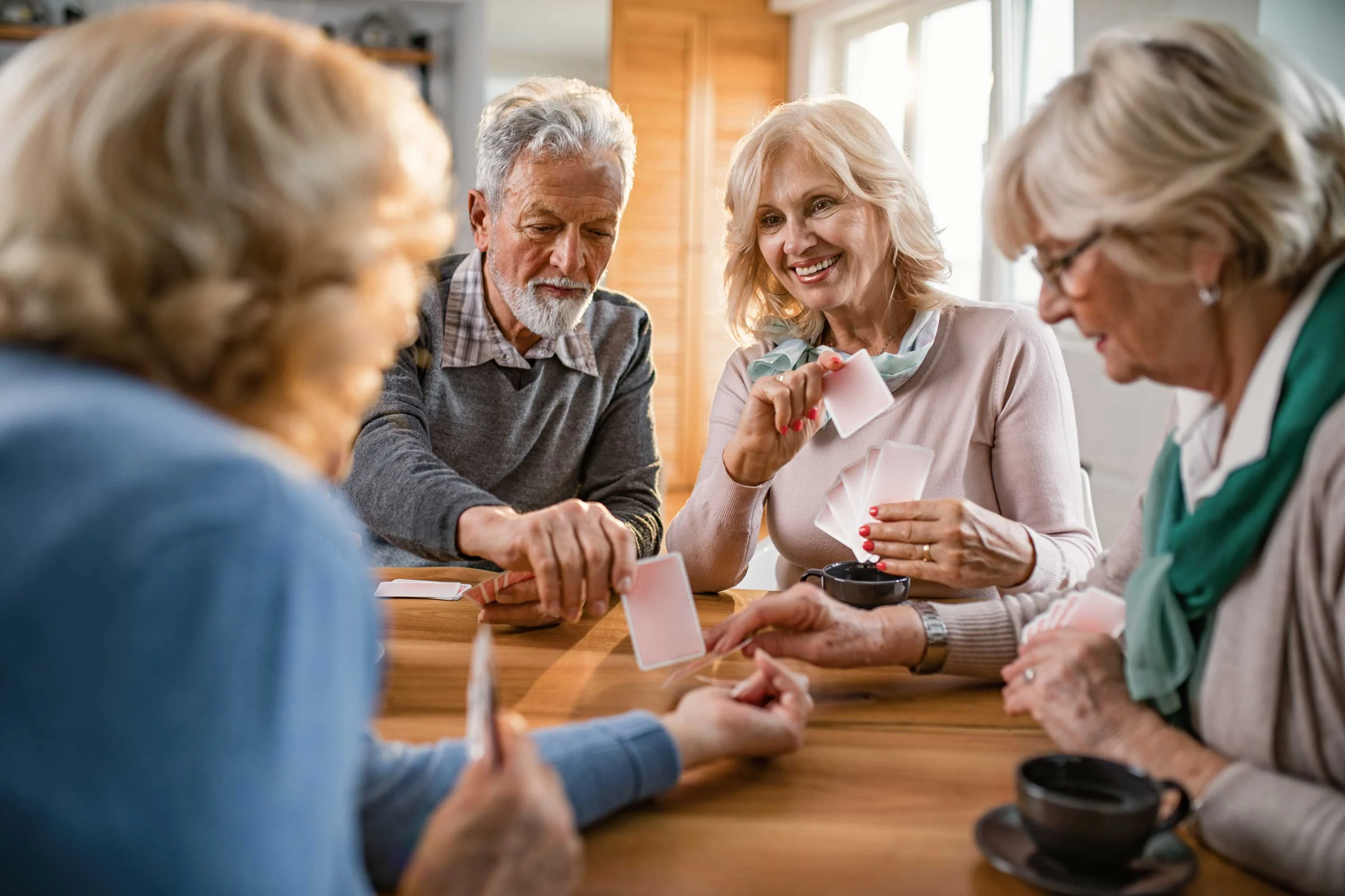 Seniors Playing Cards Together A group of elderly people sitting around a table, engaging in a card game. They are smiling and interacting with each other, with cups of coffee in front of them.